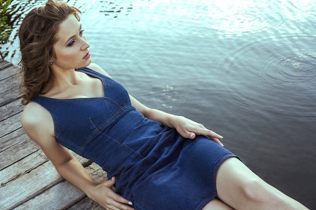Beautiful young fashion model with freckles on her face and denim blue dress and fashion makeup and hairstyle is lying down on pier, posing and looking into distance.
