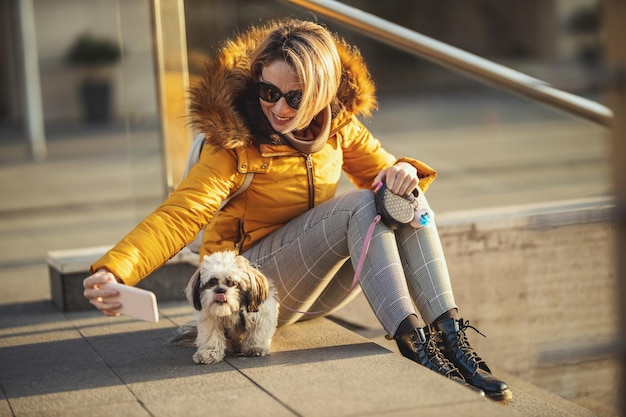 美しい若いファッションの女性は、街の通りで遊んでいる彼女のかわいいペットの犬と一緒に時間を過ごして、自分撮りをしています。