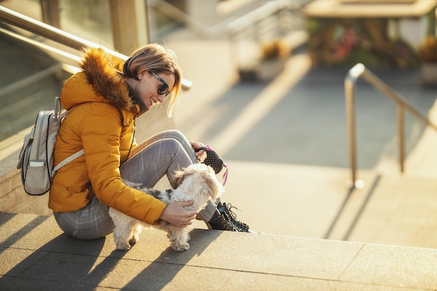 美しい若いファッションの女性は、街の通りで遊んでいる彼女のかわいいペットの犬と一緒に時間を過ごしています。