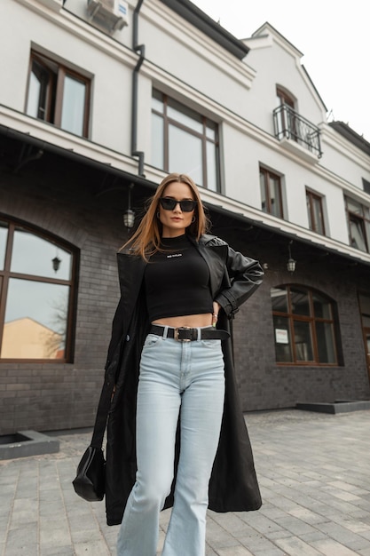 Premium Photo  Beautiful young fashionable woman in urban fashion outfit  with a long leather coat black top and high waisted jeans with a purse is  walking outside the building