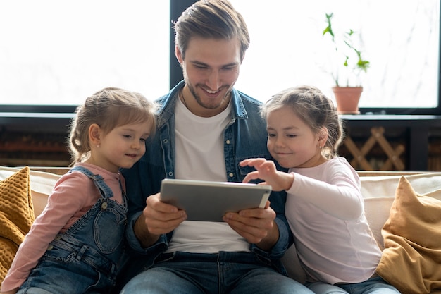 Beautiful young father, his cute little daughters are using a tablet and smiling, sitting on sofa at home.