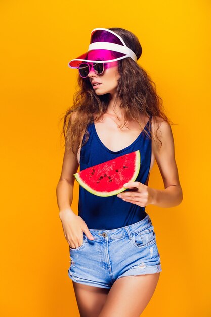 Beautiful young female in cap, sunglasses and swimsuit hold slice of watermelon and looking away while standing on yellow. Copyspase