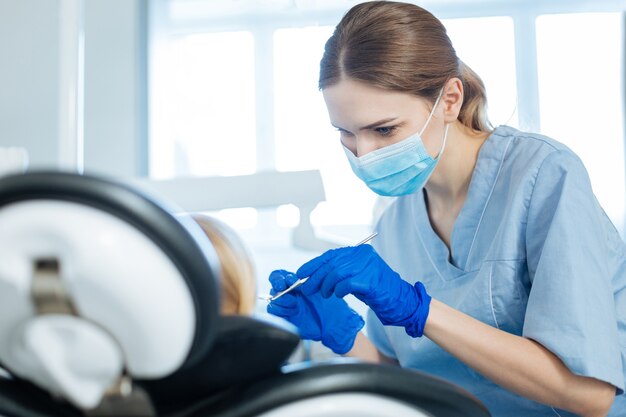 Bella giovane femmina dentista utilizzando uno specchio della bocca e controllando abilmente una cavità orale del suo piccolo paziente