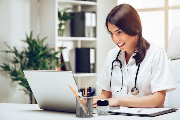 Beautiful young female doctor sitting in her consulting room, smiling and using laptop.