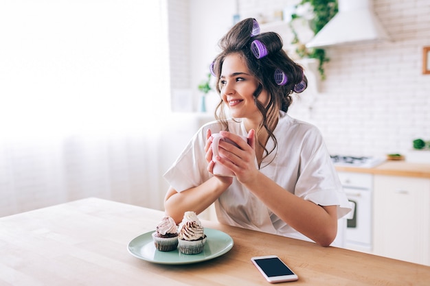 Bella giovane casalinga femminile in cucina. tenendo la tazza e guardare al lato. sorridere da solo. pancakes e telefono sul tavolo. giovane donna spensierata che vive vita ricca di beatufiul. nessun lavoro.