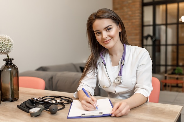 Beautiful young female medical doctor is looking at camera and smiling while writing a recipe.