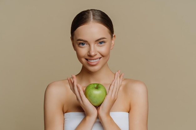 Фото Красивая молодая женщина-модель с идеальной свежей гладкой кожей держит зеленое яблоко, содержащее много витаминов, стоит в помещении, завернутая в банное полотенце, изолированное на коричневом фоне натуральная косметика