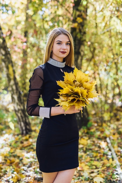 Beautiful young female student in black dress holding a bouquet of leaves in autumn park.