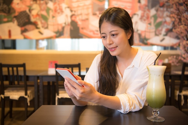 beautiful young female tourist sitting in retro style coffee restaurant leisurely using mobile phone sharing popular iced matcha green tea milk.