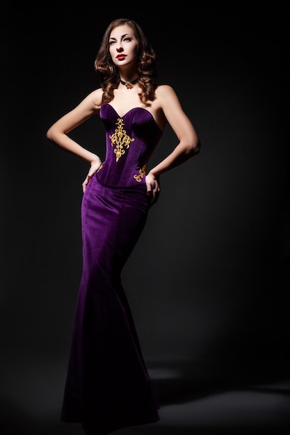 Beautiful young female wearing purple dress holding her waist. Vertical studio shot.