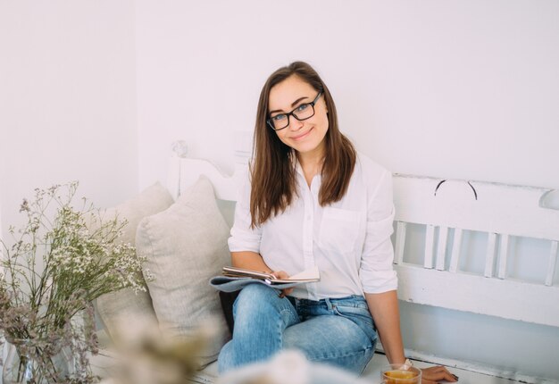 Beautiful young feminine woman in stylish glasses dressed in fashionable casual clothes sits on a bench with a notebook and a cup of tea