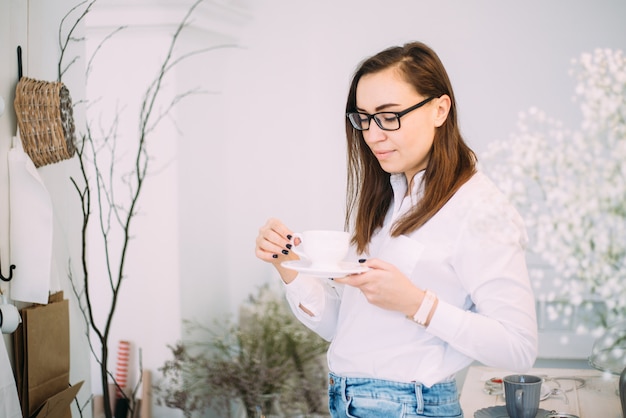 Beautiful young feminine woman in stylish glasses smiling with a cup of coffee, dressed in fashionable casual clothes in a bright room with fresh flowers