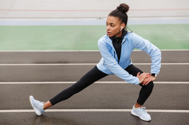 Bella giovane sportiva africana in forma che indossa auricolari wireless facendo esercizi di stretching allo stadio