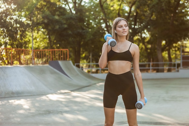 Beautiful young fit woman doing morning exercise with dumbbells outdoor.