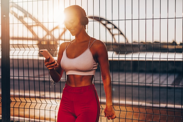 La bella donna giovane e in forma in buona forma si esercita da sola sulla strada del ponte della città. ascolta musica con cuffie e smartphone. bellissimo tramonto sullo sfondo.