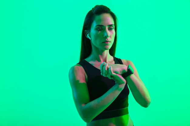Beautiful young fitness girl wearing wireless earphones isolated over green neon wall, doing stretching exercises