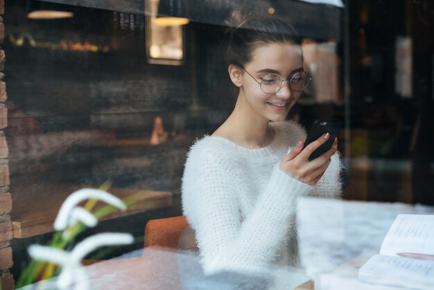 Beautiful young freelancer girl in white jacket and glasses looking into smartphone and smiling