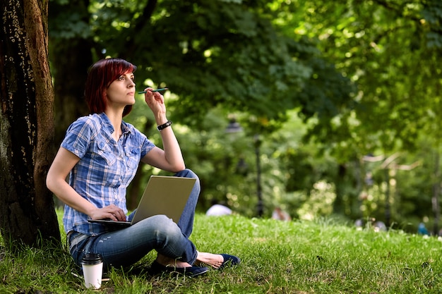 Beautiful young freelancer woman using laptop sitting under the tree in the park.