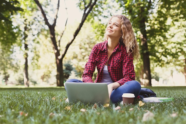 Beautiful young freelancer working on laptop and drinking coffee, sitting on the grass outdoors in the park. Technology, communication, education and remote working concept, copy space