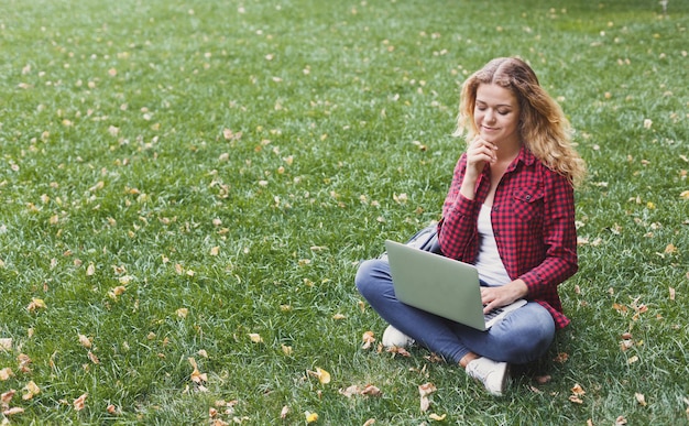 Beautiful young freelancer working on laptop, sitting on the grass in a pleasant atmosphere outdoors in the park. Technology, communication, education and remote working concept, copy space