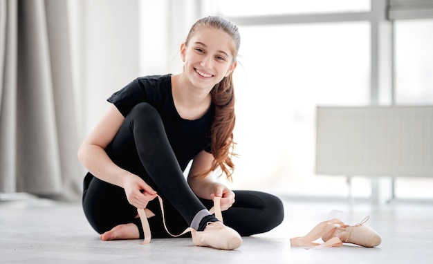 Ballerina di bella ragazza in abito nero seduto sul pavimento, sorridendo alla telecamera e indossando scarpe da punta