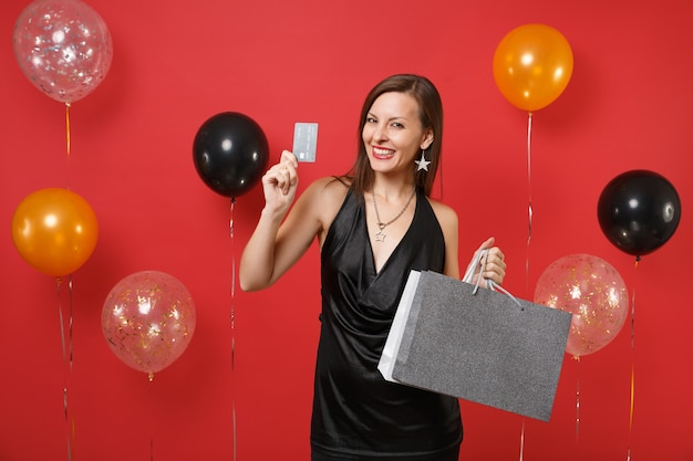 Beautiful young girl in black dress hold credit card, colored packages bags with purchases after shopping on bright red background air balloons. Happy New Year, birthday mockup holiday party concept.