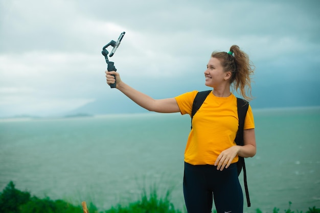 Beautiful young girl blogger outdoors walking traveling hiking holding stabilizer with smartphone recording video vlog waving to camera smiling woman doing a selfie on top of a mountain