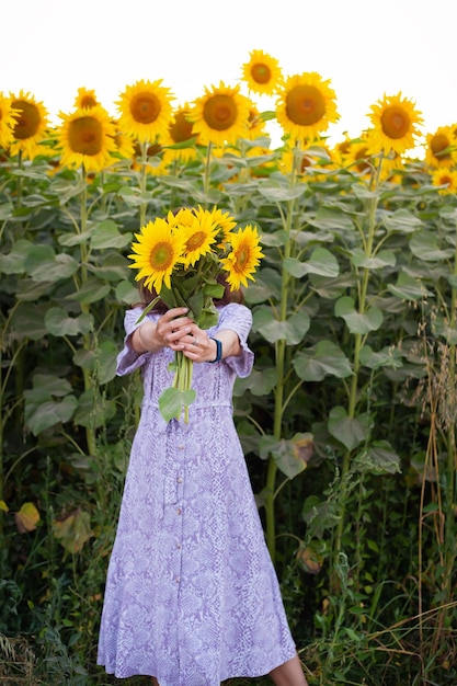 A beautiful young girl in a dress holds a bouquet of sunflowers\
in her hands stands on the road among a large field with\
sunflowers