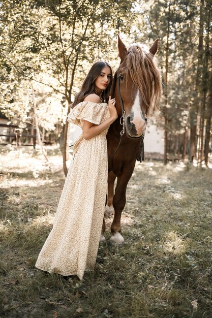 A beautiful young girl dressed in a dress stands near a horse in the forest