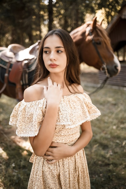 A beautiful young girl dressed in a dress stands near a horse in the forest