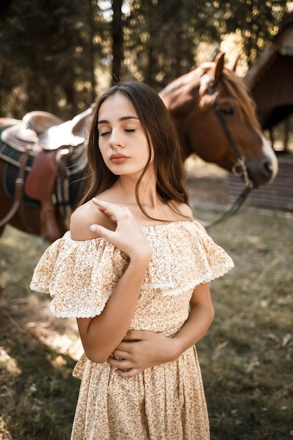 A beautiful young girl dressed in a dress stands near a horse in the forest