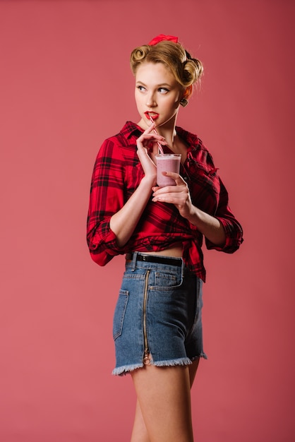 Beautiful young girl dressed in retro style holding a milkshake on the pink background. Pinup portrait