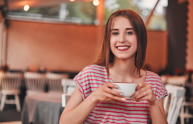 Beautiful young girl drinks cappuccino while relaxing in cozy cafe and smiling