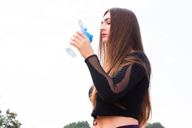 美しい少女は運動後、公園で水を飲みます。ジョギング後の公園で水を飲むスポーティな女性の肖像画。トレーニングトレーニング後の女性アスリート。コピースペース