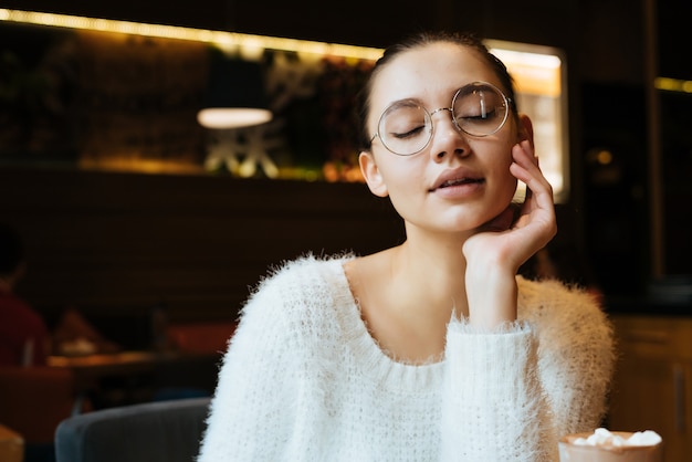 Beautiful young girl freelancer with glasses sitting in a cafe, thinking about work, closed her eyes