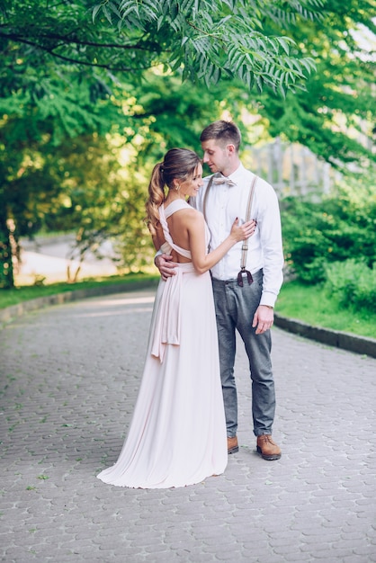 Beautiful young girl in a gently pink dress hugged to her boyfriend while walking in the park at sunset