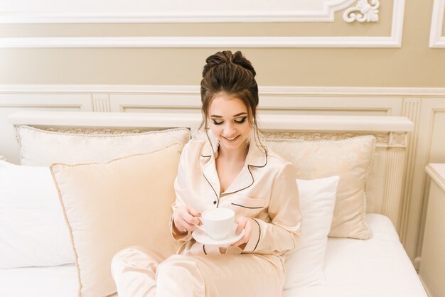 세련 된 머리와 화장 커피 한 잔 함께 침대에 누워 고급스러운 방에 황금 잠 옷에서 아름 다운 젊은 여자. 아침 신부.