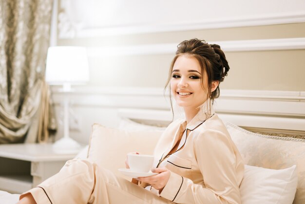 Beautiful young girl in Golden pajamas in a luxurious room with stylish hair and makeup lying in bed with a Cup of coffee. Morning bride.