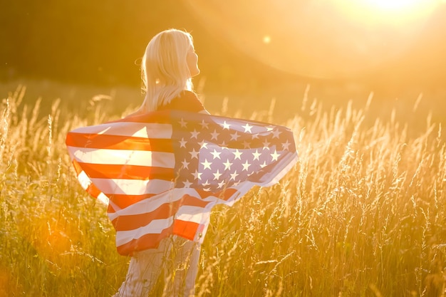 Beautiful young girl holding an American flag in the wind in a field of rye. Summer landscape against the blue sky. Horizontal orientation.