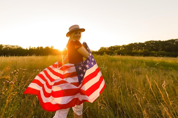 ライ麦畑で風にアメリカの国旗を保持している美しい少女。青い空を背景にした夏の風景。水平方向。