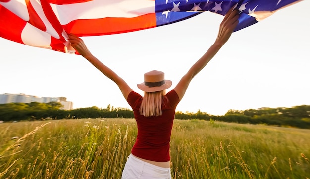 Beautiful young girl holding an American flag in the wind in a field of rye. Summer landscape against the blue sky. Horizontal orientation.