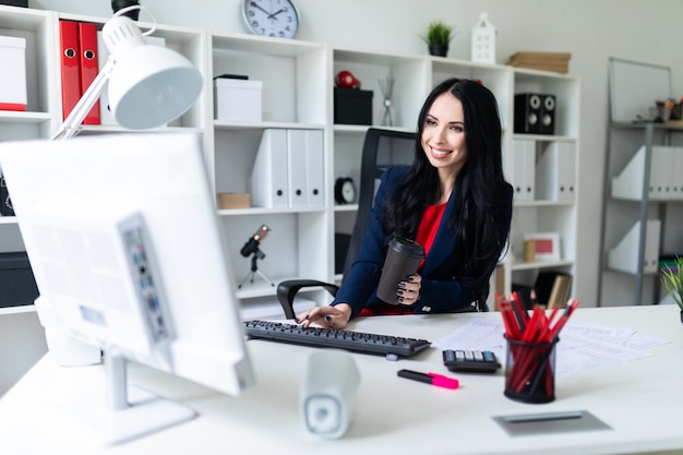 Beautiful young girl holding a glass in her hand with coffee and typing text on the keyboard