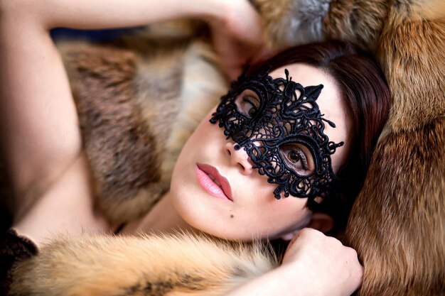 Photo a beautiful young girl in a lacy black mask lies on a red fur