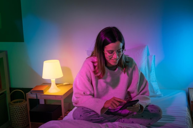 Bella ragazza che guarda il suo telefono sul letto con la stanza dei colori al neon tecnologia a letto concept