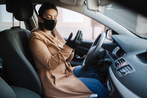 Beautiful young girl in a mask sitting in a car and put on protective gloves.