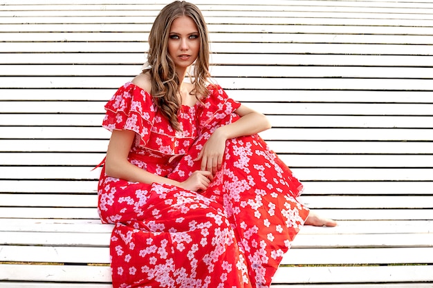 Beautiful young girl, model, brown-haired woman with long hair in a red dress sits on a bench and poses with a relaxed, languid look.
