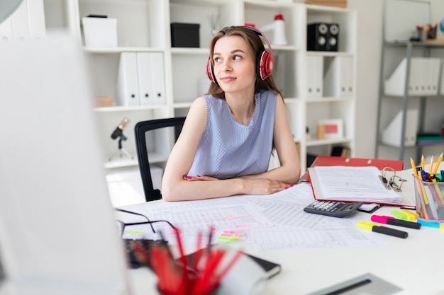 Beautiful young girl in the office sits at a table in red headphones.