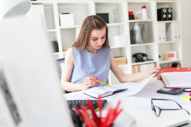 Bella ragazza in ufficio si siede a un tavolo e lavora con una matita, un blocco note e documenti.