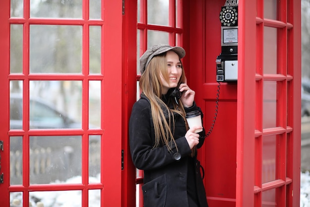 Beautiful young girl in a phone booth. The girl is talking on the phone from the payphone. English telephone booth in the street and a woman talking on the phone.