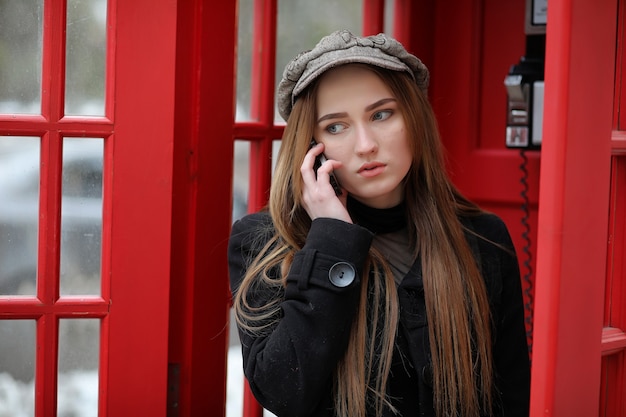 Beautiful young girl in a phone booth. The girl is talking on the phone from the payphone. English telephone booth in the street and a woman talking on the phone.